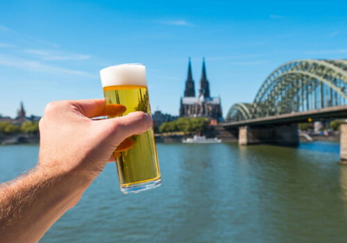 Eine Hand hält ein Glas Kölsch vor die Kamera. Im Hintergrund sind der Rhein und der Kölner Dom zu sehen.